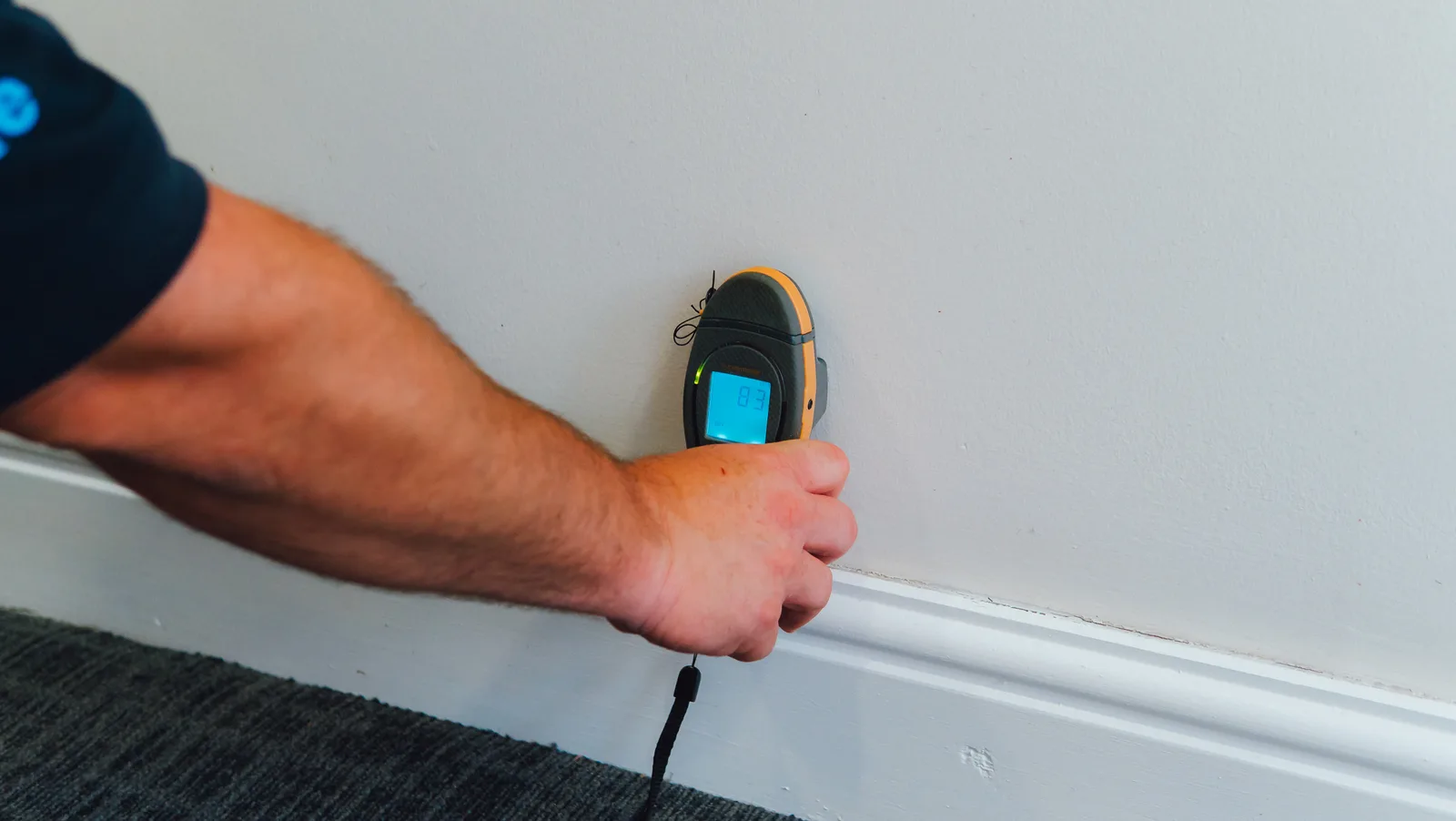 An engineer holding a pinless moisture meter against a wall above a skirting board, the display showing a high reading